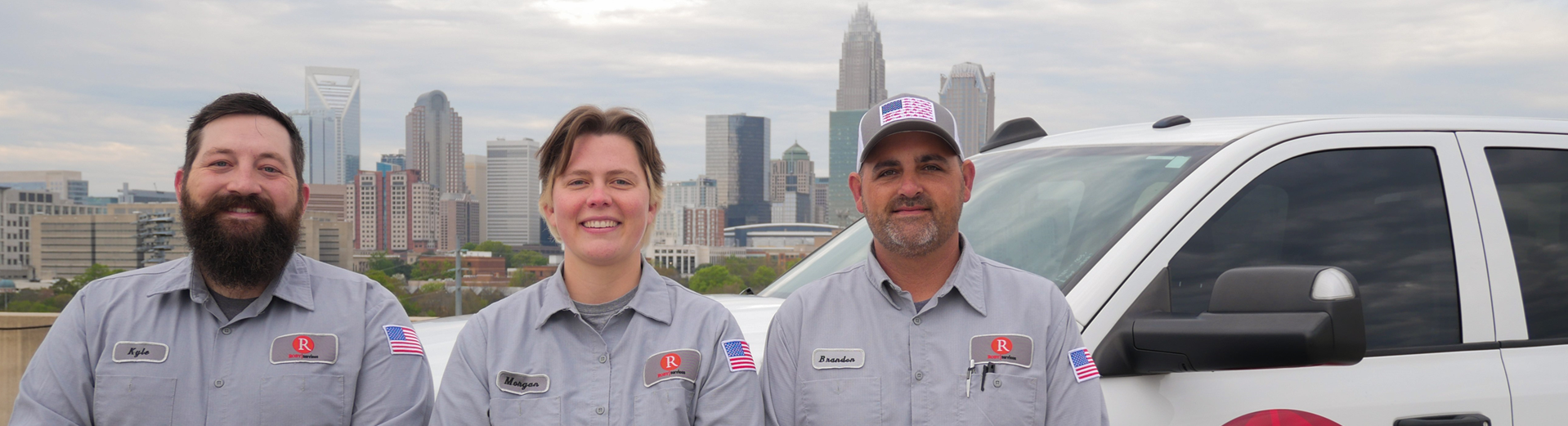 Three uniformed technicians stand in front of a company truck with a red "R" logo, with a city skyline in the background on a cloudy day.